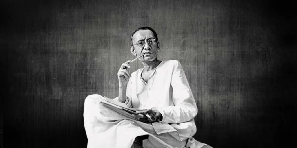 A black and white portrait of Saadat Hasan Manto seated cross-legged in a white kurta, a pen raised to his lips and papers on his lap
